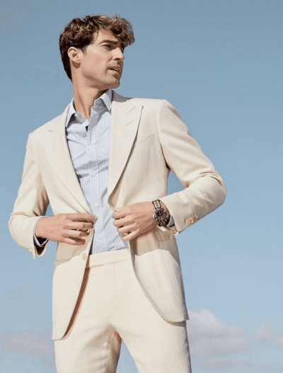 KURZ - Uhrenmarken - Stilvoll gekleideter Herr in einem crème-farbigen Anzug trägt eine Omega Uhr der Speedmaster Kollektion. Im Hintergrund sieht man den Himmel.